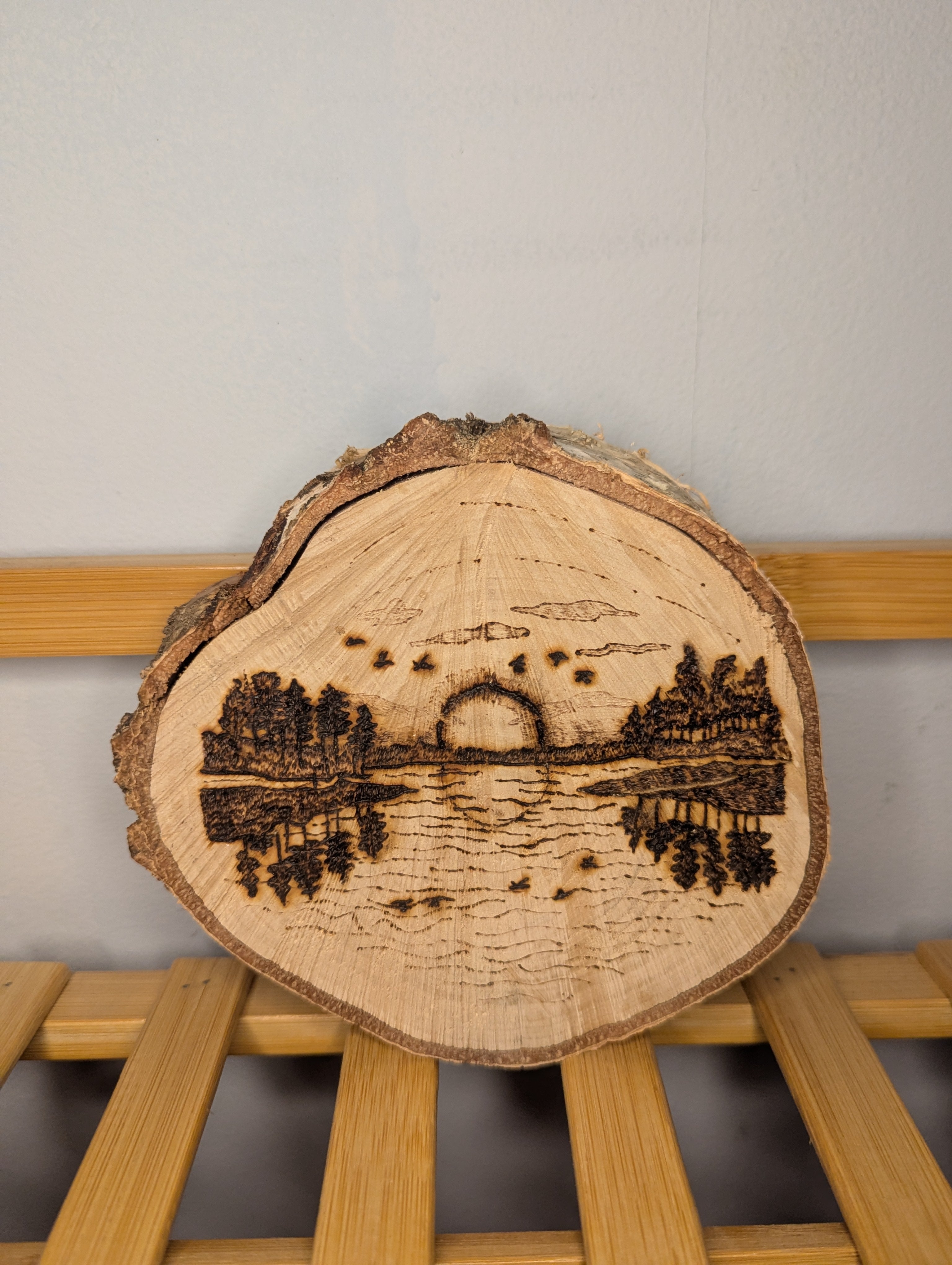 Hand-burned pyrography of a lakeside sunset with pine trees reflected in the water on a natural round live-edge wood slice