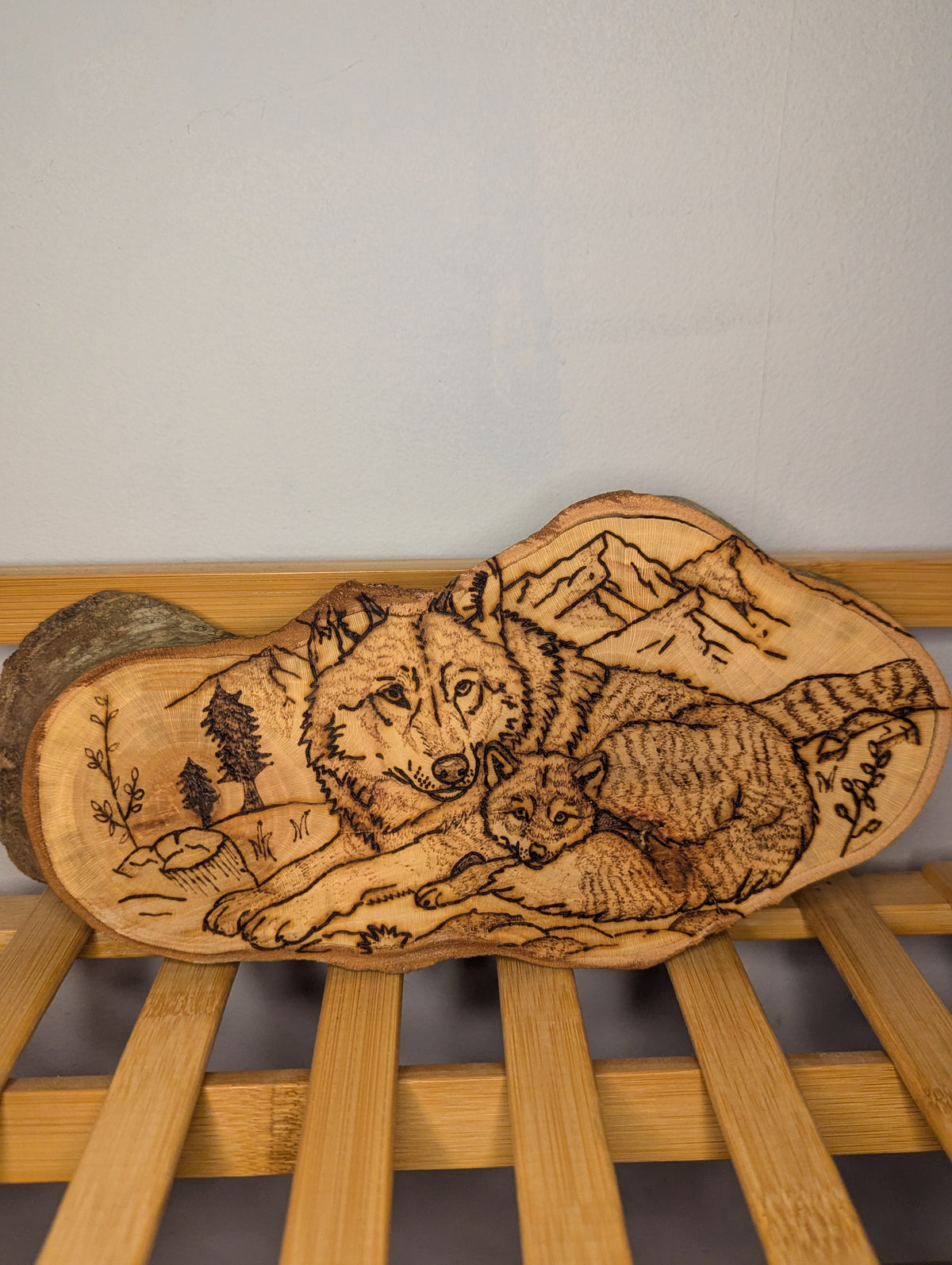 Hand-burned pyrography of a wolf and cub on a natural live-edge wood slice with mountain and forest background